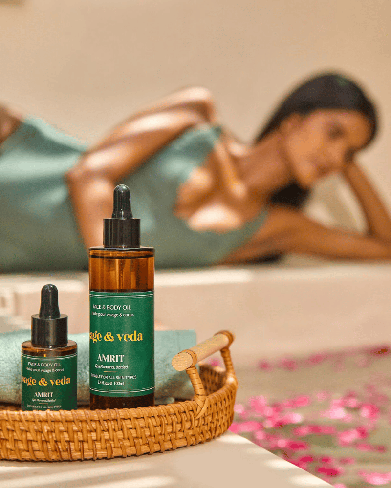 Lifestyle image of woman sitting by the pool, two Sage & Veda Amrit Face & Body Oil products shown in focus